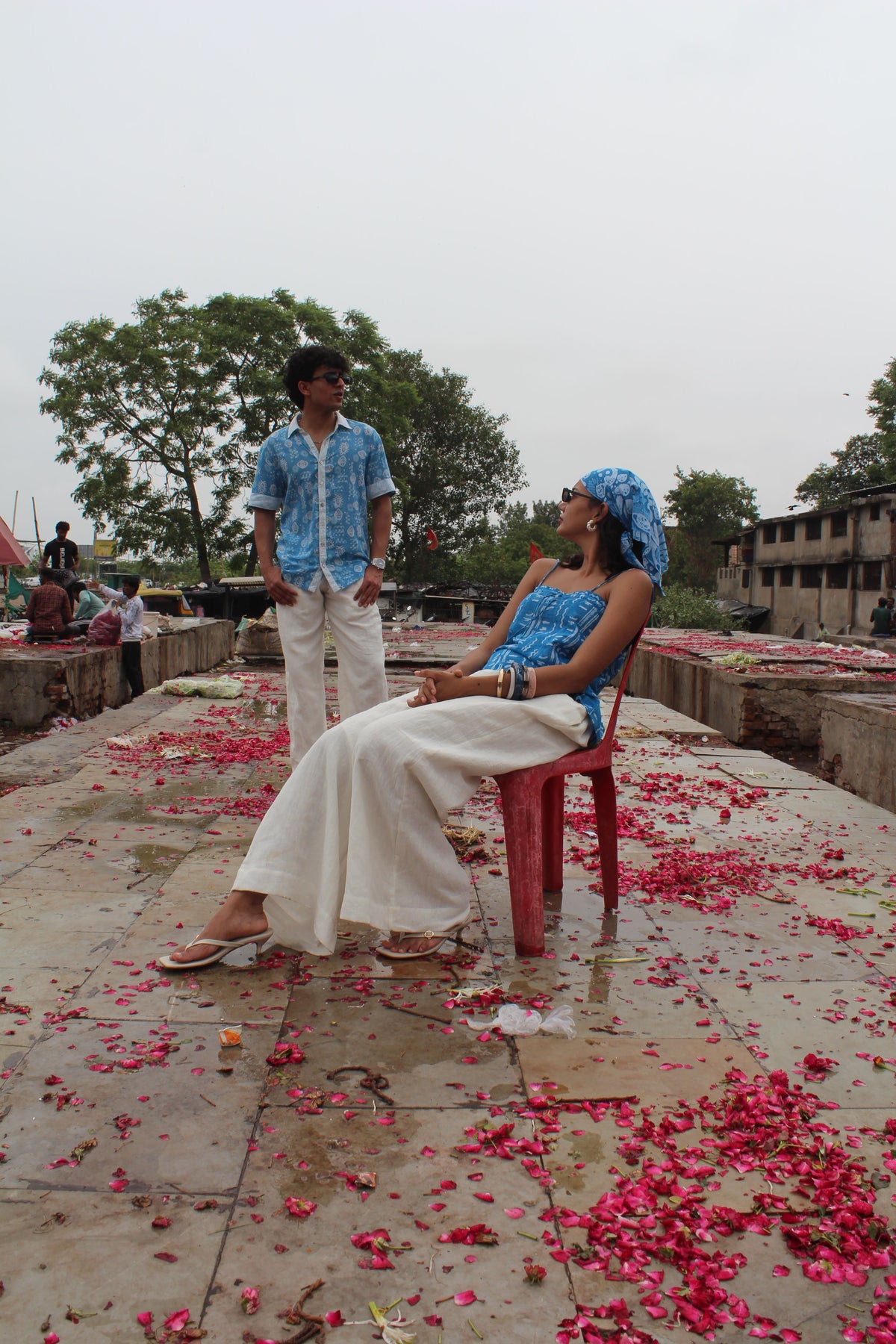 A man and a woman in traditional Indian attire pose outdoors on a stone platform scattered with pink flower petals. The woman wears a white saree and blue headwrap, sitting on a red plastic chair, while the man in a blue patterned shirt and white trousers stands behind her.